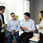 Professional team consulting on internet while discussing ongoing project. Four employees talking in meeting workspace, using gadgets and drinking coffee. Interaction concept